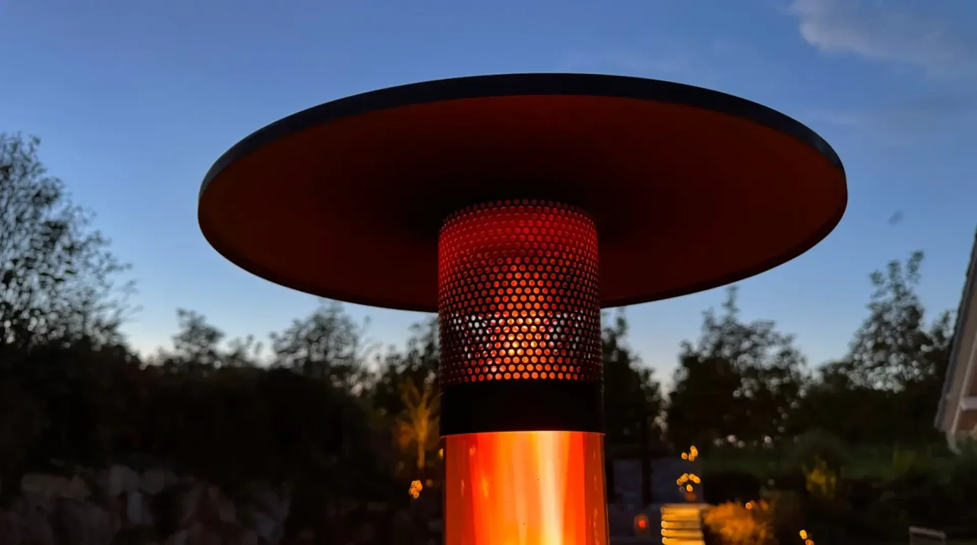 Eine Nahaufnahme eines Terrassenheizers, der in der Abenddämmerung orange leuchtet, mit Bäumen, die sich im Hintergrund gegen den Abendhimmel abheben.