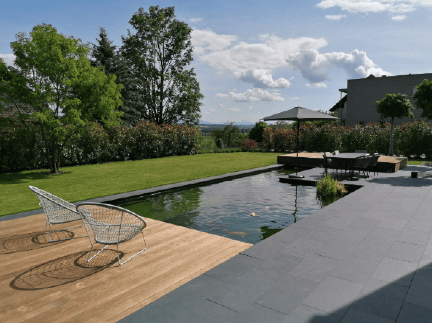 Modern backyard with a rectangular pond, wooden deck with two wire chairs, gray stone patio, outdoor dining table under a large umbrella, and lush green landscaping under a partly cloudy sky.