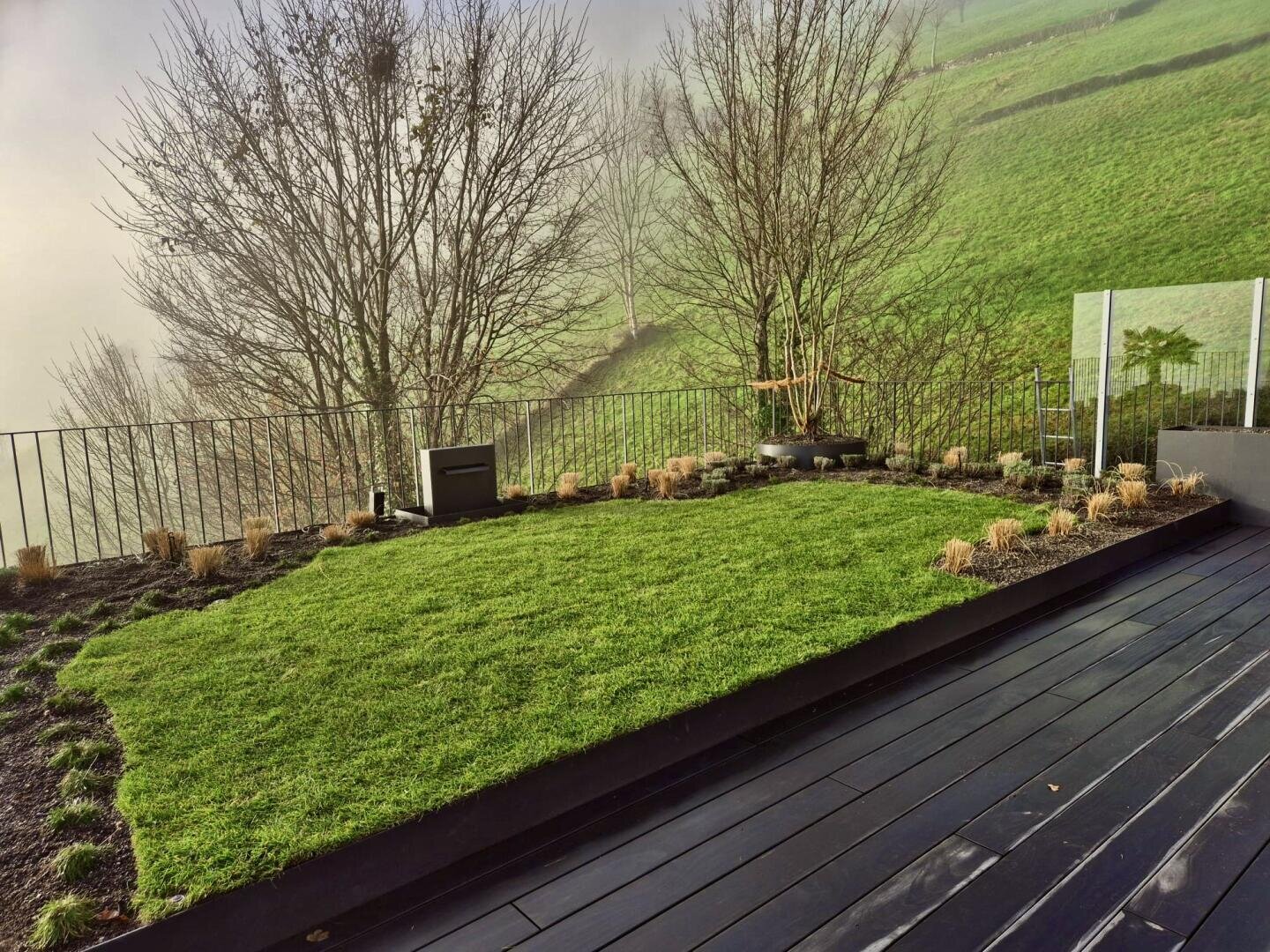 Ein kleiner, ordentlich gemähter Rasen mit umliegenden Gartenbeeten liegt neben einer dunklen Holzterrasse mit Blick auf einen nebligen Hügel mit kahlen Bäumen und saftig grünem Gras, der von einem Metallgeländer und Glasscheiben begrenzt wird.