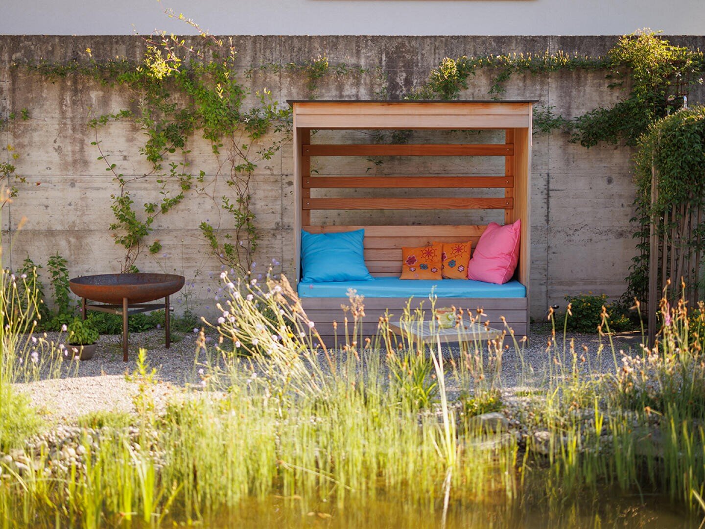 Eine gemütliche hölzerne Gartenbank mit bunten Kissen steht an einer mit Weinreben bewachsenen Betonwand. In der Nähe befindet sich eine runde Feuerstelle, mit hohen Gräsern und einem kleinen Teich im Vordergrund.