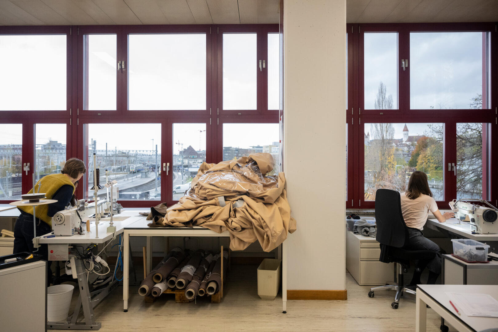 In einem hellen Raum mit großen Fenstern arbeiten zwei Personen an Nähmaschinen. Ein Stapel beiger Stoffe und Stoffrollen liegen auf einem Tisch zwischen ihnen. Draußen sind Gebäude, Bäume und Eisenbahnschienen zu sehen.