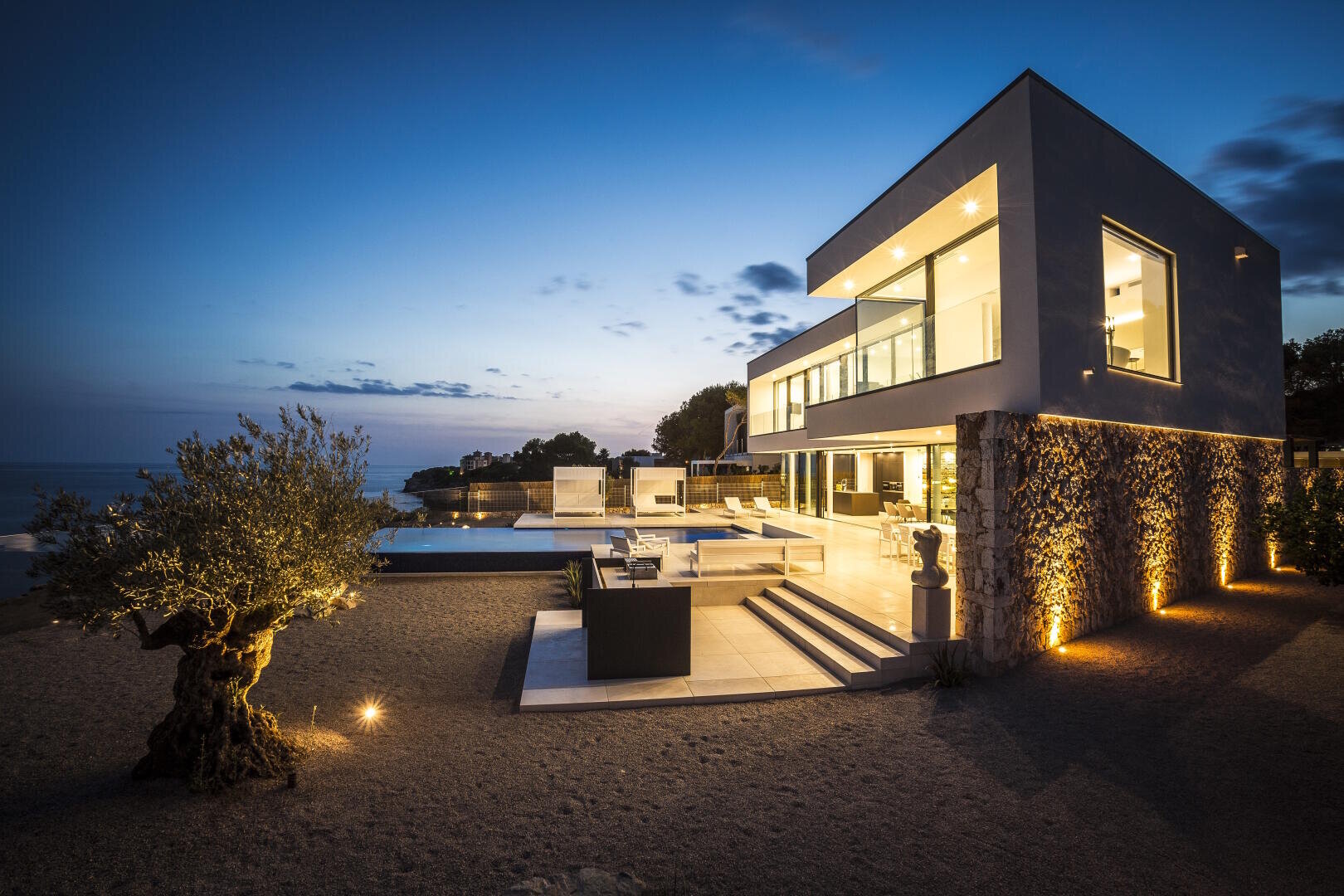 Modern house with large windows and outdoor terrace, illuminated at dusk, overlooking a calm sea. The scene is characterized by minimalist landscaping with a tree and decorative lights that accentuate the architecture.
