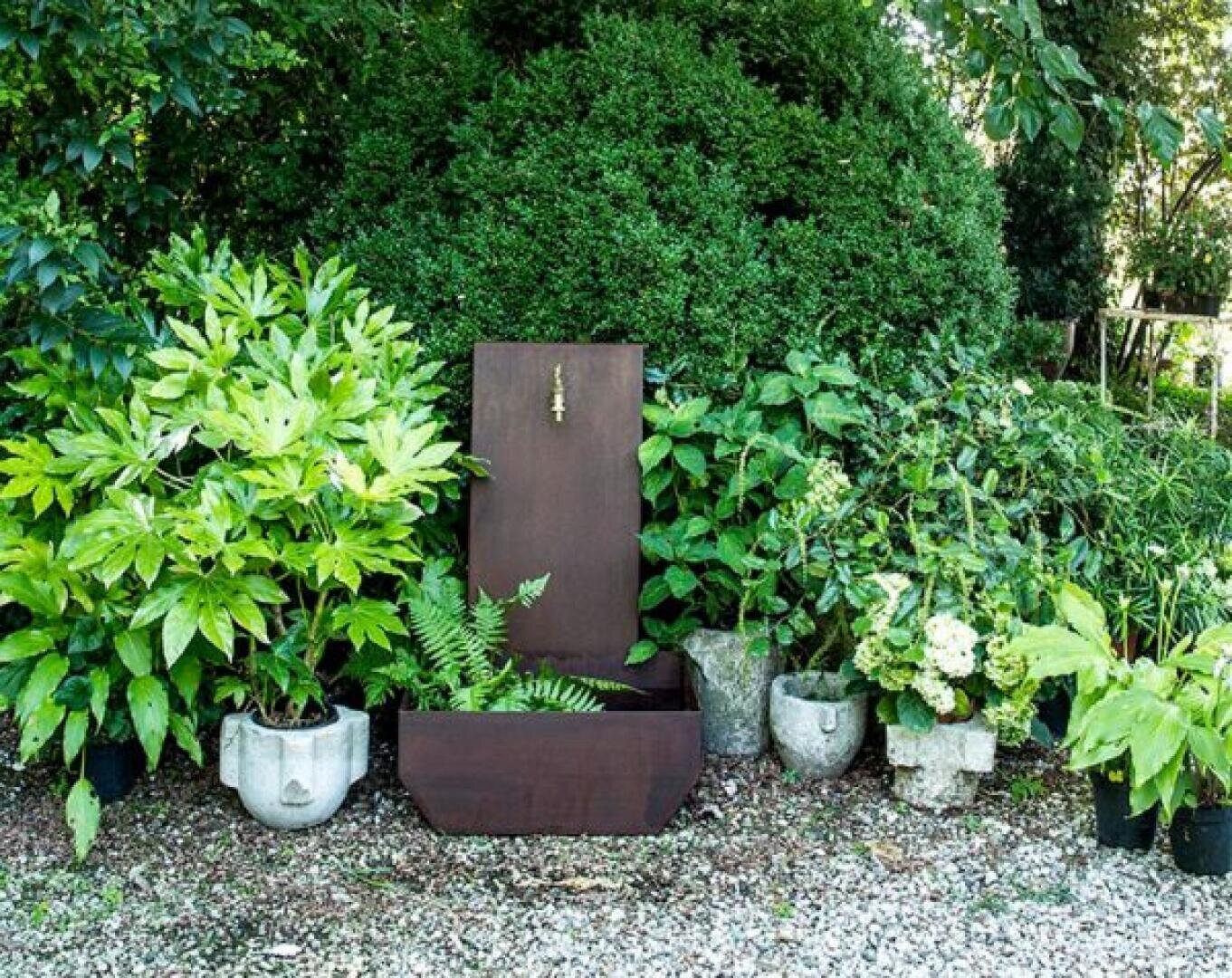 A rustic brown metal garden trough with its lid open, revealing ferns inside, is surrounded by various green leafy plants and shrubs in pots, set on gravel in a lush, outdoor garden.