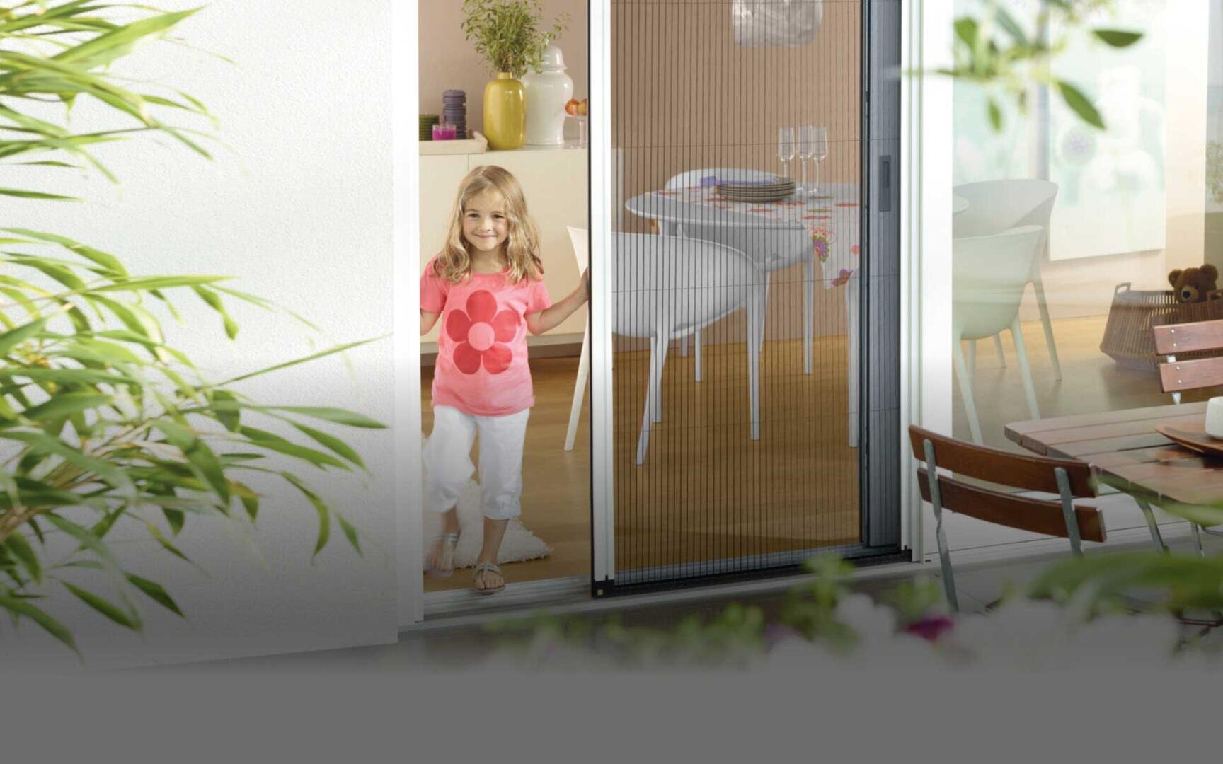 Ein junges Mädchen, das ein rosa Hemd mit einer großen Blume trägt, steht an einer Schiebetür, lächelt und hält den Griff fest. Im Innenraum sind ein Esstisch und Stühle zu sehen, draußen stehen Pflanzen und Terrassenmöbel.