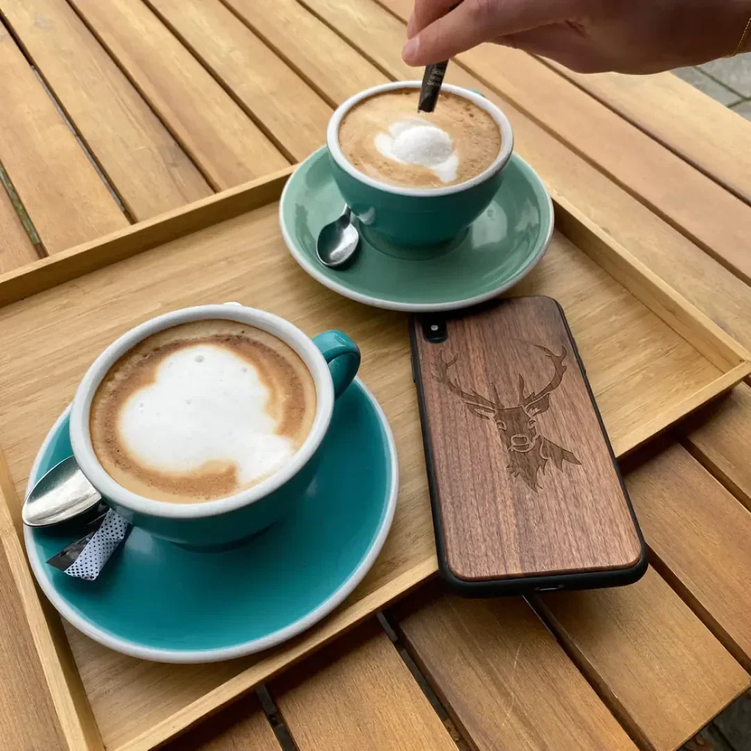 Two cups of cappuccino in blue-green mugs on a wooden tray, one of them stirred, with spoons and sugar packets next to it. On the tray is a telephone with a wooden case engraved with a stag.