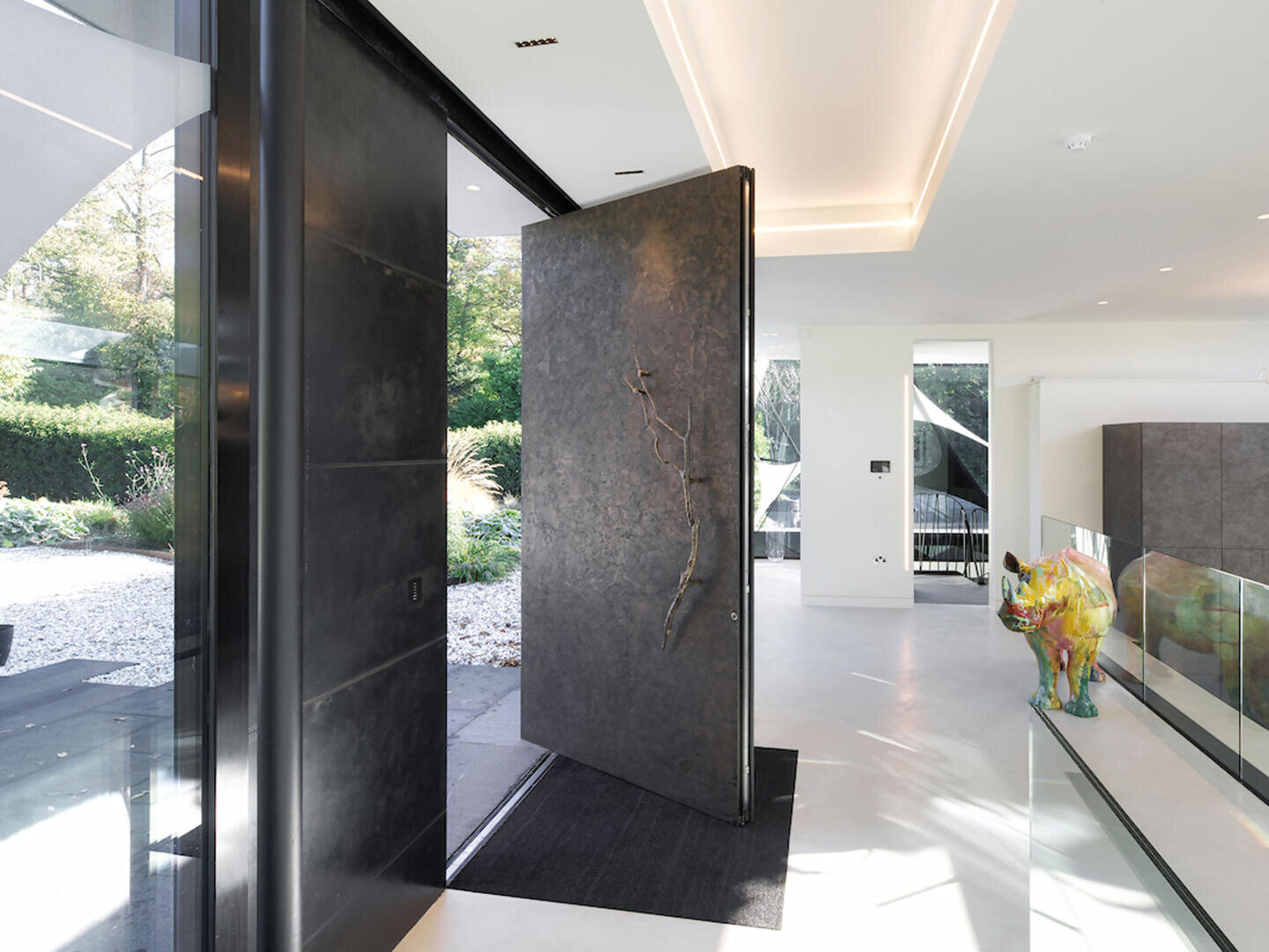Modern entrance with a large, dark revolving door that is partially open and offers a view of a landscaped garden. The interior is characterized by plain white walls, minimal decor and a colourful animal sculpture on the right-hand side.