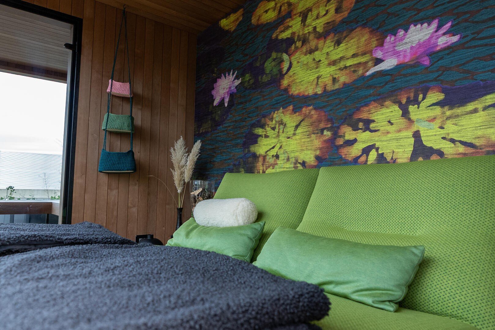 A cozy bedroom with a green bed, green pillows and a white bolster pillow. There is a colorful mural of water lilies on the wooden wall and a window and hanging baskets on the left side. A soft gray blanket covers the bed.