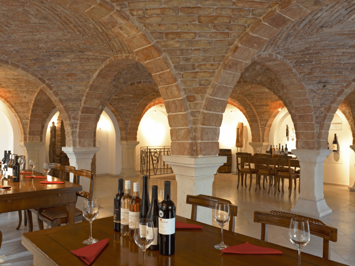 Ein Weinkeller mit gewölbten Ziegeldecken, Holztischen mit Weinflaschen, Gläsern und roten Servietten. Die warme Beleuchtung unterstreicht die Steinsäulen und die gemütliche, rustikale Atmosphäre.