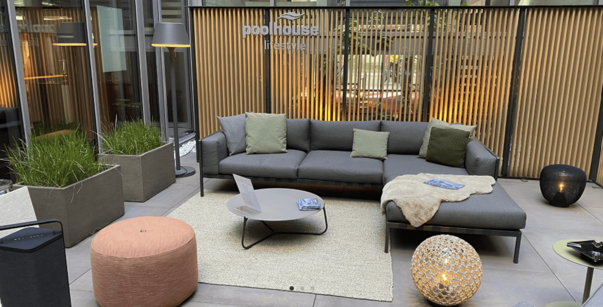 Moderne Outdoor-Lounge mit einem grauen Sektionssofa, grünen und beigen Kissen, einer hellbraunen Decke, einem runden Couchtisch, einer strukturierten Leuchte, einem pfirsichfarbenen Pouf, hohen Pflanzgefäßen und vertikalen Holzlatten als Hintergrund. Poolhouse Lifestyle ist auf Glas geschrieben.