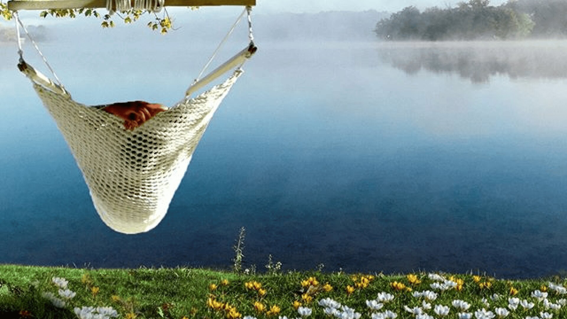 Eine Person entspannt sich in einer weißen Hängematte, die über Gras und Blumen an einem ruhigen See hängt, während im Hintergrund Nebel aus dem Wasser aufsteigt und Bäume zu sehen sind.
