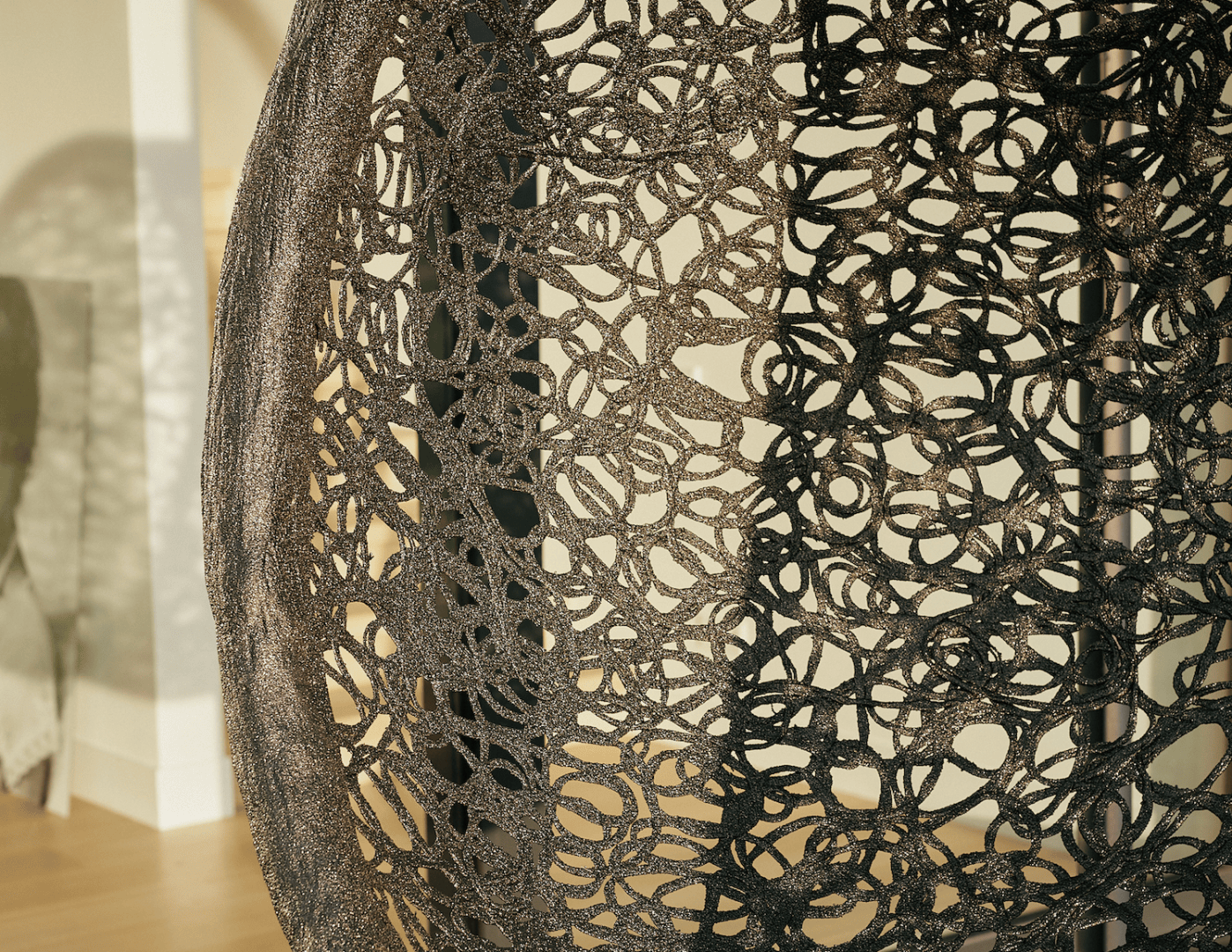 Nahaufnahme eines geflochtenen Stuhls oder Dekorationsobjekts aus dunklem, geschlungenem Material mit einem komplizierten, offenen, spitzenartigen Muster, das Schatten auf eine sonnenbeschienene Wand und einen Holzboden im Hintergrund wirft.