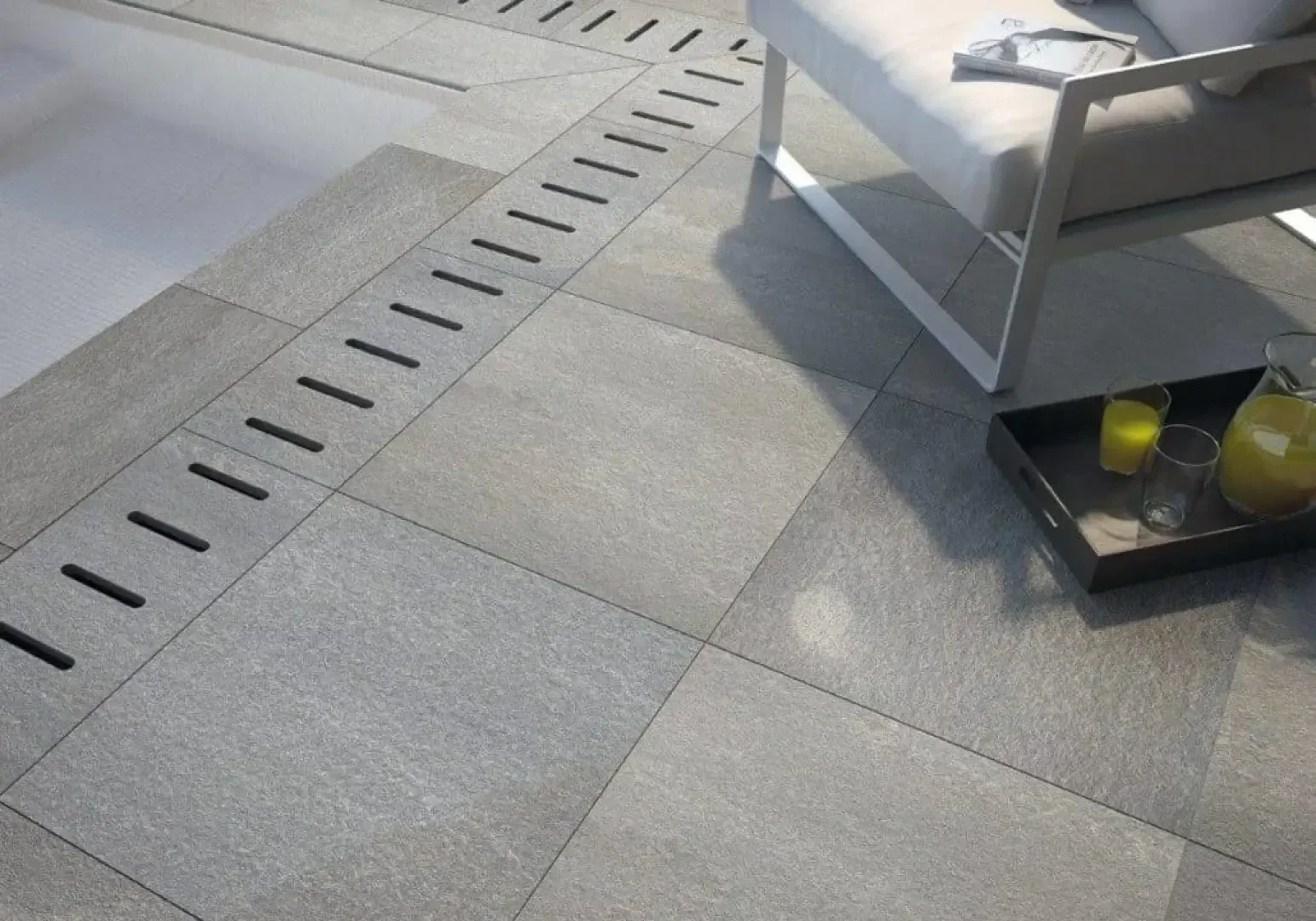 Modern outdoor patio with large gray stone tiles, a linear metal drainage grate, a white cushioned chair, and a tray holding a glass pitcher of juice and two glasses.