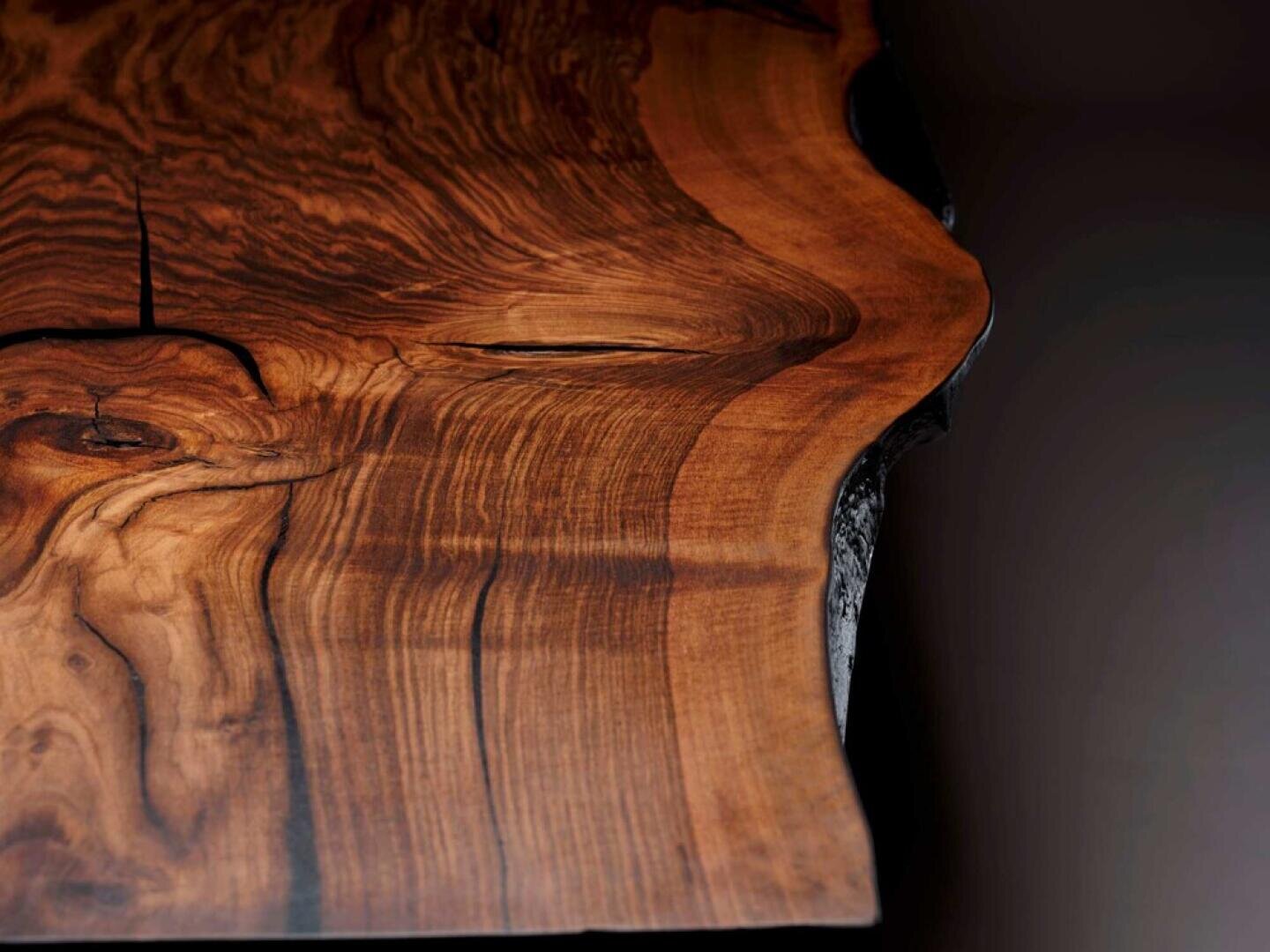 Close-up of a polished wooden slab with rich, dark and light brown grain patterns and a natural, uneven edge, set against a dark background.