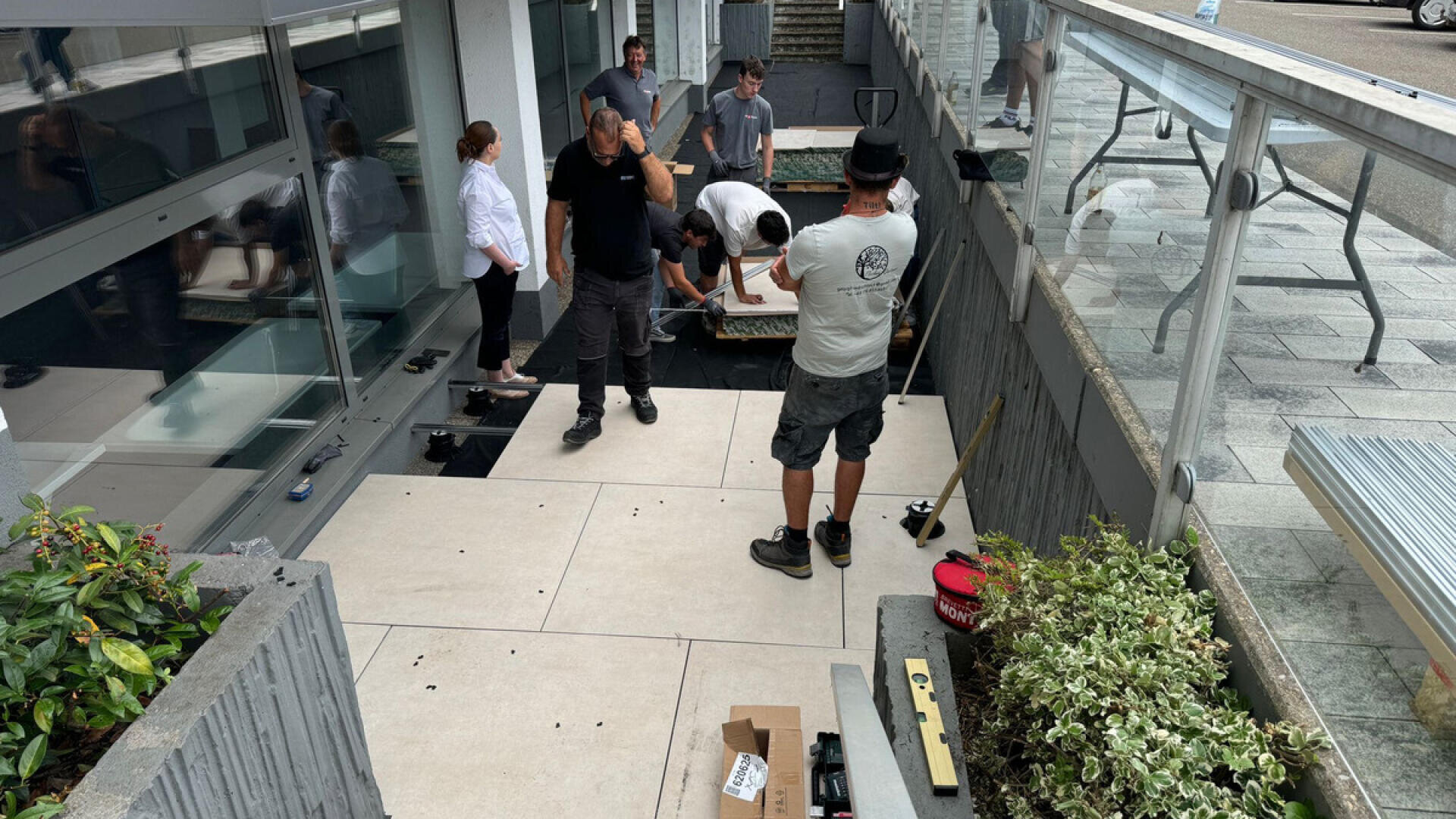 A group of people work together installing large beige tiles on an outdoor patio surrounded by glass and concrete barriers, with tools and materials scattered around. Some people are actively working while others observe.