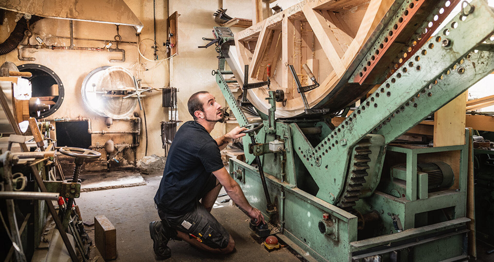 Ein Mann kniet neben einer großen, grünen Industriemaschine in einer Werkstatt und justiert deren Steuerung. Holzteile und mechanische Komponenten umgeben ihn, und Sonnenlicht strömt durch ein rundes Fenster herein.