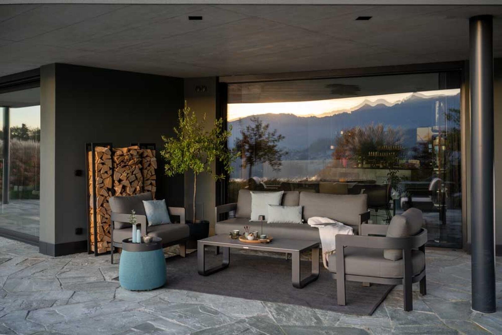 Moderne Außenterrasse mit grauen Polstermöbeln, einem Couchtisch mit Getränken und Snacks, einem Stapel Brennholz, einer Topfpflanze und großen Glasfenstern, in denen sich die Berge bei Sonnenuntergang spiegeln.
