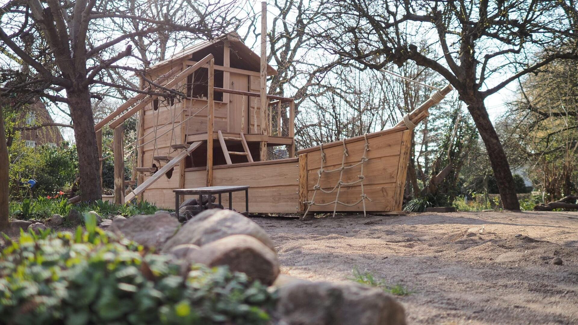 Ein hölzernes Spielgerät in Form eines Schiffes mit einer Strickleiter, einer Treppe und einem Aussichtsturm steht zwischen Bäumen in einem sandigen Außenbereich. Im Vordergrund sind Felsen und Grünpflanzen zu sehen.