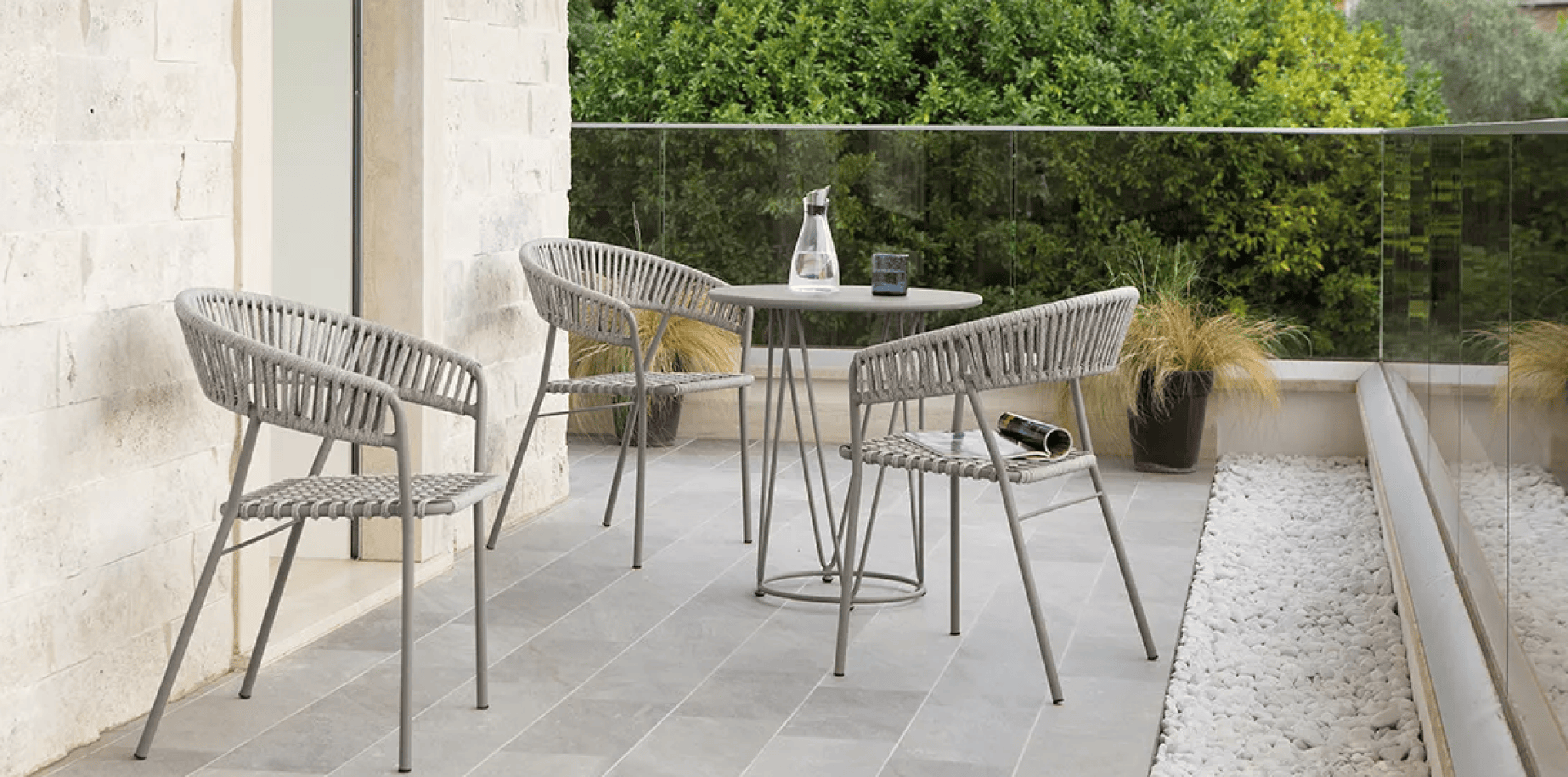 Eine moderne Terrasse mit drei geflochtenen Stühlen und einem runden Tisch mit einer Glaswasserflasche und Bechern. Der Raum ist mit grauen Fliesen, weißen Steinen, Topfpflanzen und einem Glasgeländer mit grünem Laub im Hintergrund ausgestattet.