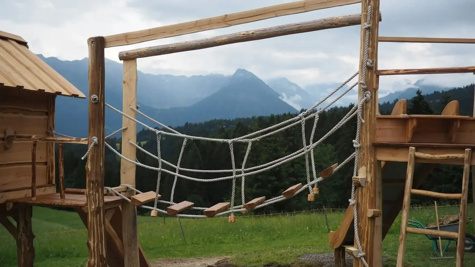 A wooden playground structure with a rope bridge and slide, set on grass with mountains and forest in the background under a cloudy sky.
