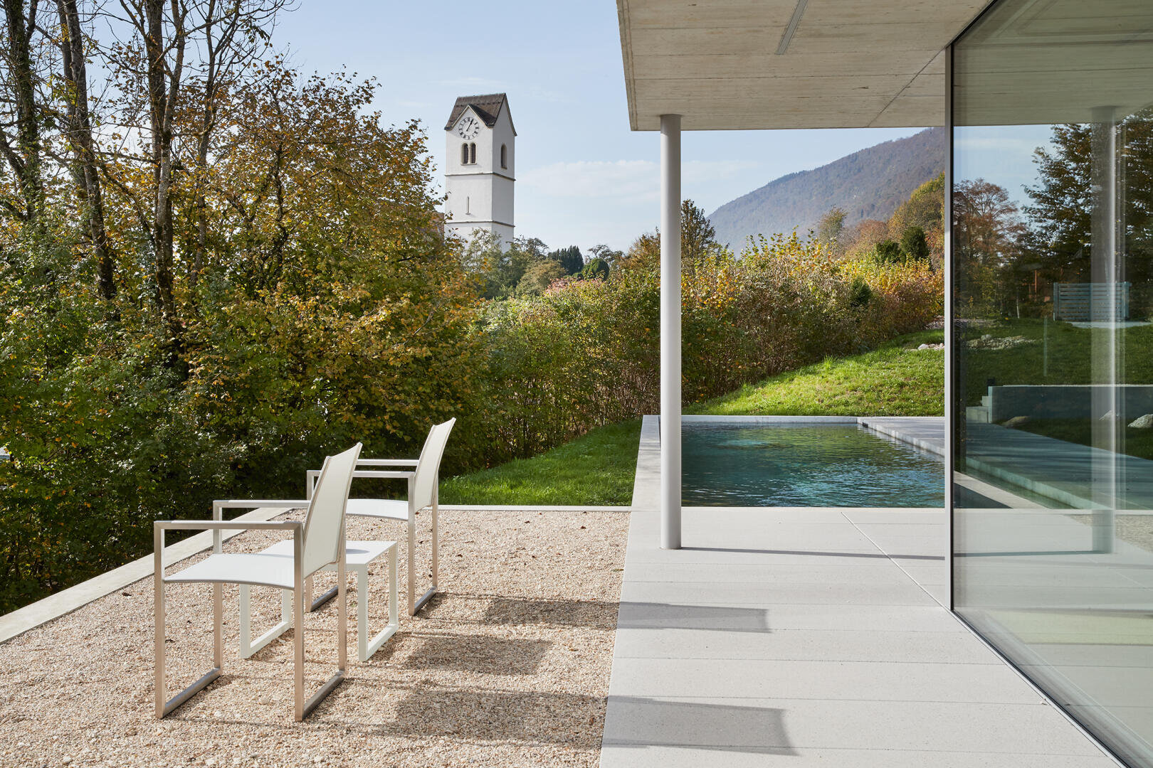 Zwei weiße Stühle stehen auf einer Kiesterrasse neben einem modernen Haus mit großen Glasfenstern, mit Blick auf einen Pool, eine grüne Landschaft und einen weißen Kirchturm in der Ferne, mit Hügeln und Bäumen im Hintergrund.