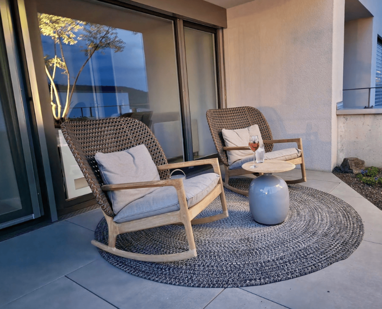 Two upholstered wicker rocking chairs with wooden armrests stand on a round woven carpet on a terrace. Between them is a small round table with two drinks. Large glass doors and part of a white building can be seen in the background.