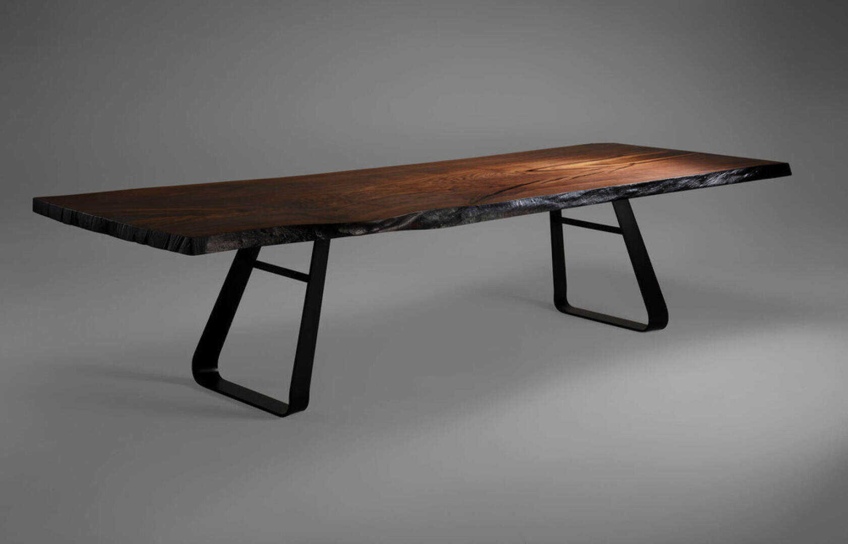 A modern table with a thick, polished wooden top with a natural, uneven edge, supported by two black metal legs with a geometric pattern, set against a plain gray background.