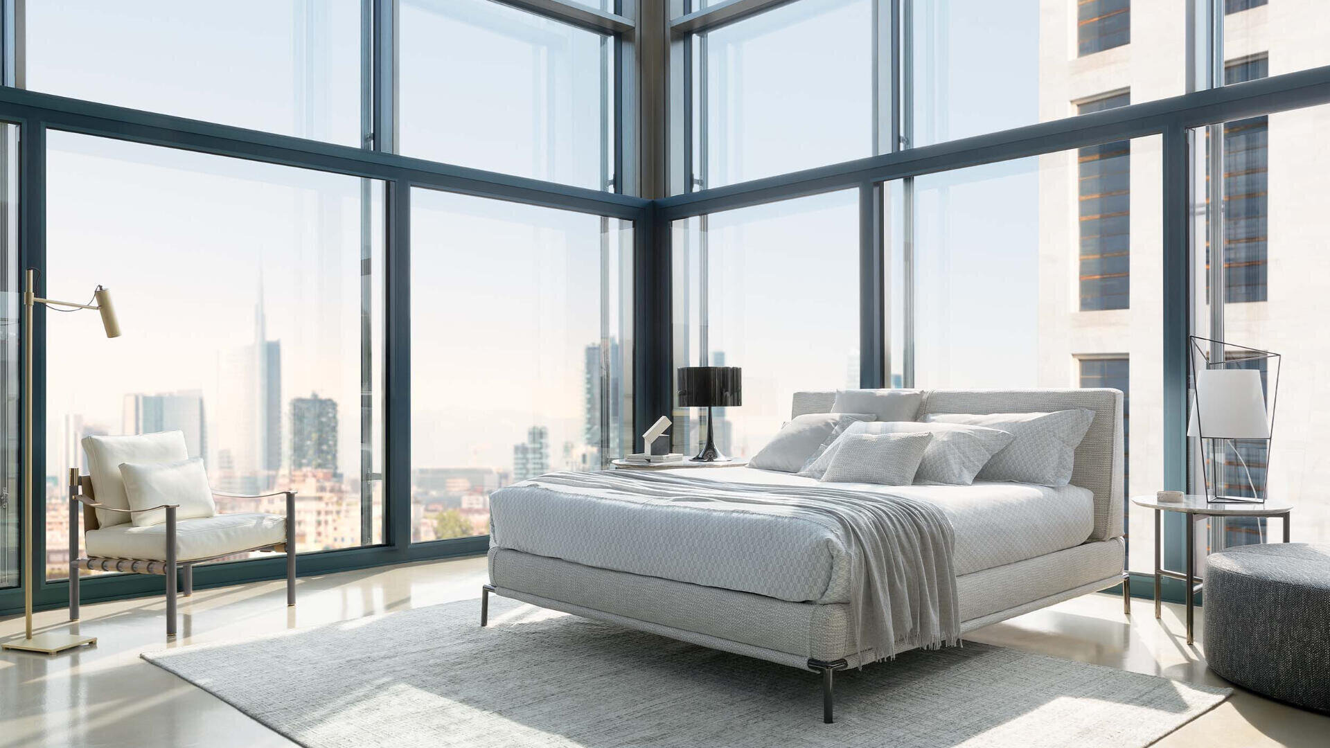 A modern bedroom with floor-to-ceiling windows and a view of a cityscape, furnished with a gray bed, white bed linen, a gray armchair, a floor lamp, a side table with lamp and a light-colored rug.