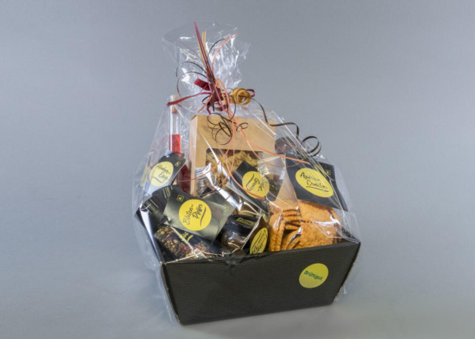 A gift basket wrapped in transparent cellophane, filled with various snacks and items, each labeled with yellow stickers. The basket is tied with a red and gold ribbon and stands against a plain gray background.