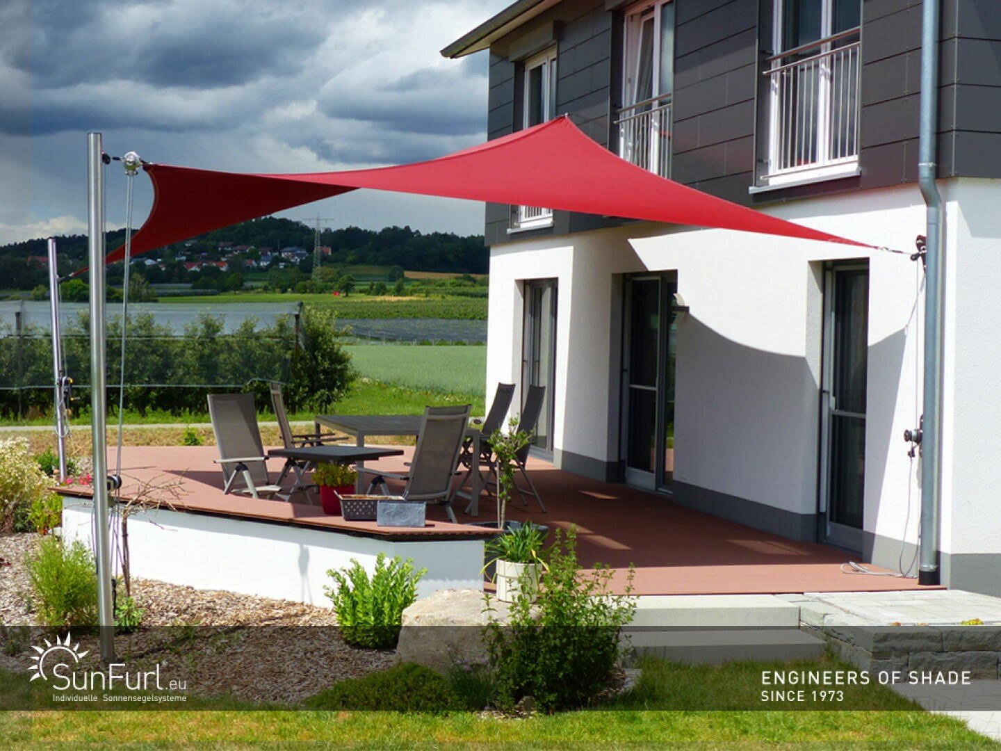 Ein rotes dreieckiges Sonnensegel ist über einer modernen Terrasse mit Sitzgelegenheiten im Freien installiert, die an ein zweistöckiges Haus angebaut ist. Der Hof ist begrünt, und im Hintergrund sind grüne Felder und Hügel zu sehen.