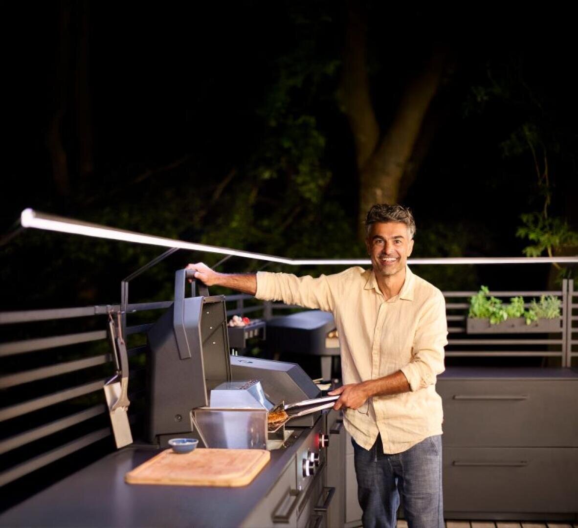 A man in a beige shirt smiles as he grills food in an outdoor kitchen at night. He lifts the lid of a grill, utensils and a wooden chopping board lie on the counter next to him. Trees and plants can be seen in the background.