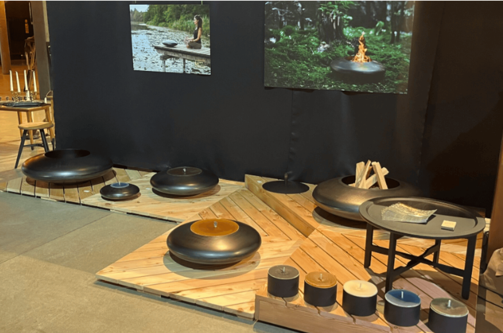A display of modern black fire pits and accessories arranged on a wooden floor, with logs, candles, and photos of fire pits in nature mounted on a black wall in the background.
