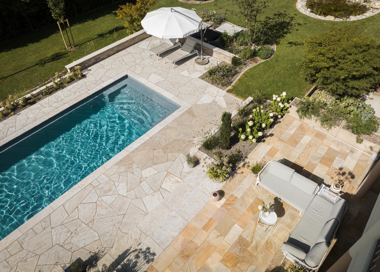 Aerial view of a backyard with a rectangular swimming pool, a stone terrace, a sofa, a table, potted plants, two deck chairs and a white parasol, surrounded by green lawn and trees.