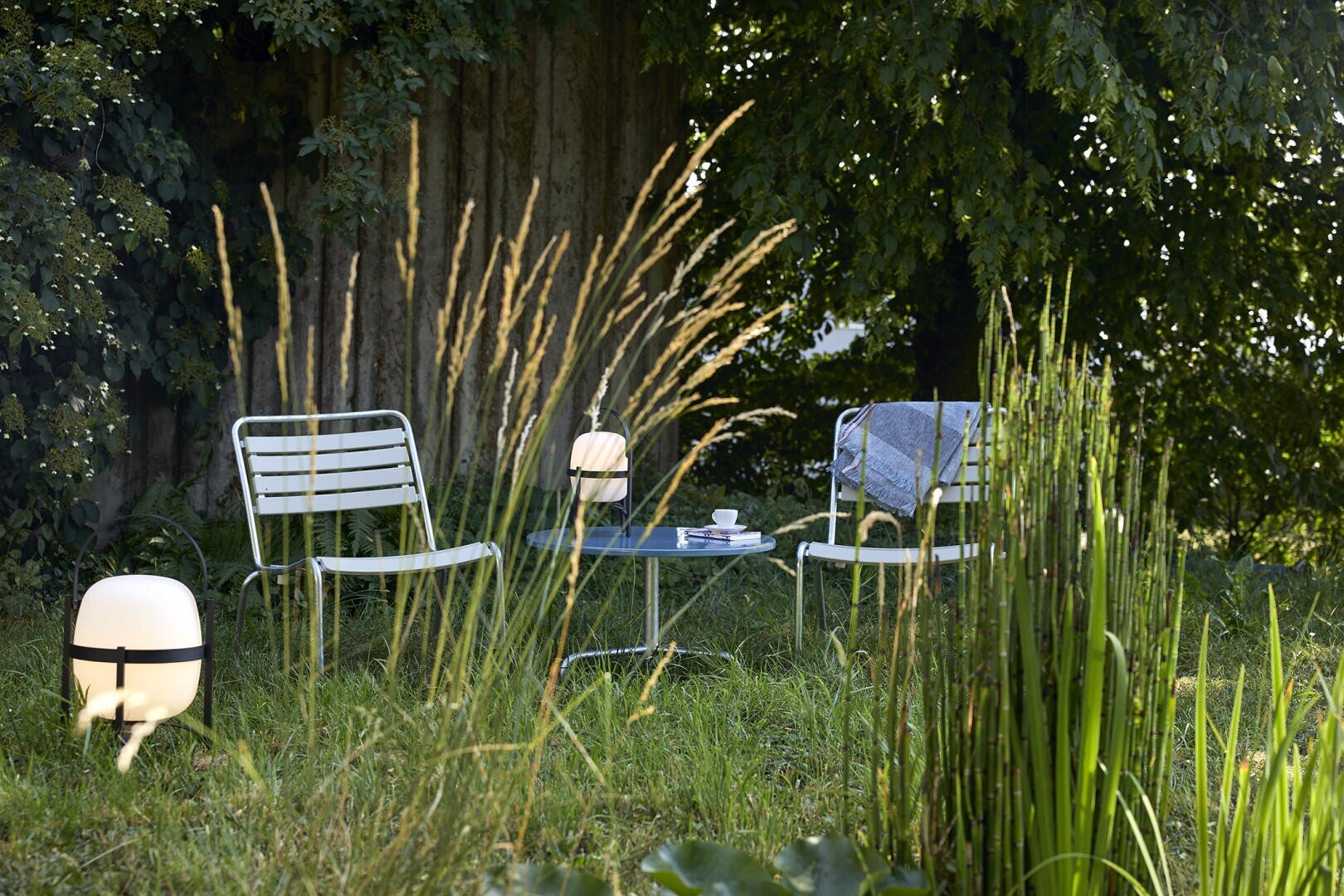 Zwei Metallstühle und ein kleiner runder Tisch mit einer Tasse und einer Lampe stehen auf dem Rasen in einem schattigen, üppigen Garten. Hohe Gräser und belaubte Bäume umgeben den gemütlichen Sitzplatz im Freien. Über einen Stuhl ist eine Decke drapiert.