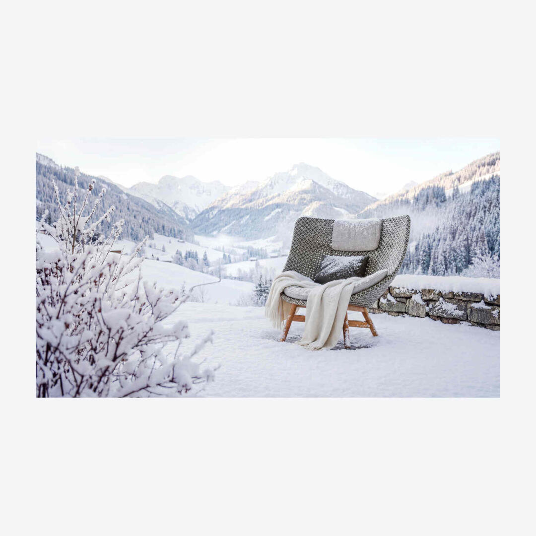 Ein gemütlicher Korbsessel mit Decke und Kissen steht auf einer verschneiten Terrasse mit Blick auf ein malerisches Wintertal und schneebedeckte Berge. Sträucher und eine Steinmauer sind unter einem wolkenverhangenen Himmel mit Schnee bedeckt.