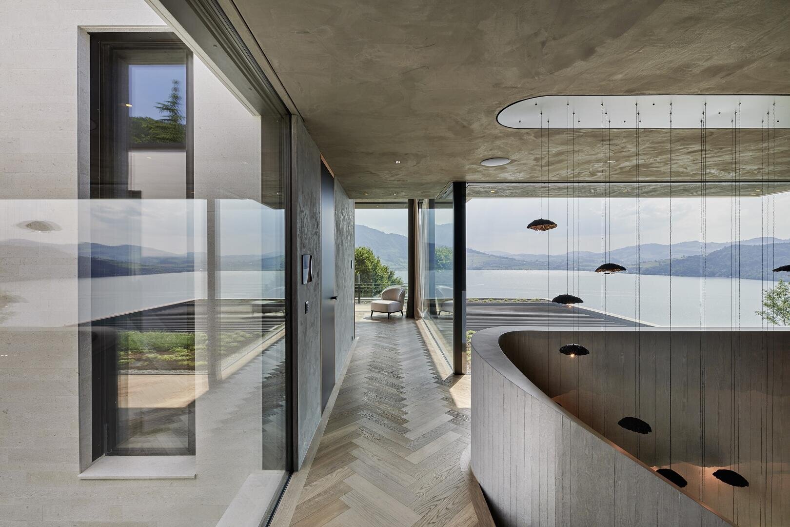 A modern interior hallway with large floor-to-ceiling windows and wooden floors offering panoramic views of a lake and the mountains. There is a curved staircase to the right and a chair by the window at the end of the hallway.