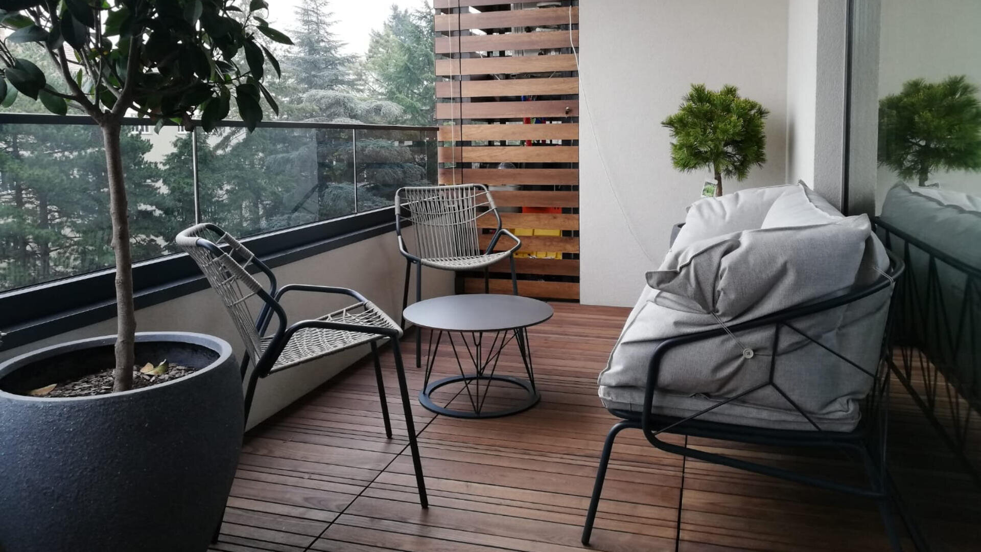Ein moderner Balkon mit Holzfußboden, zwei Metallstühlen, einem kleinen runden Tisch, einem gepolsterten Loungesessel und Topfpflanzen. Draußen sind ein Glasgeländer und Bäume zu sehen.