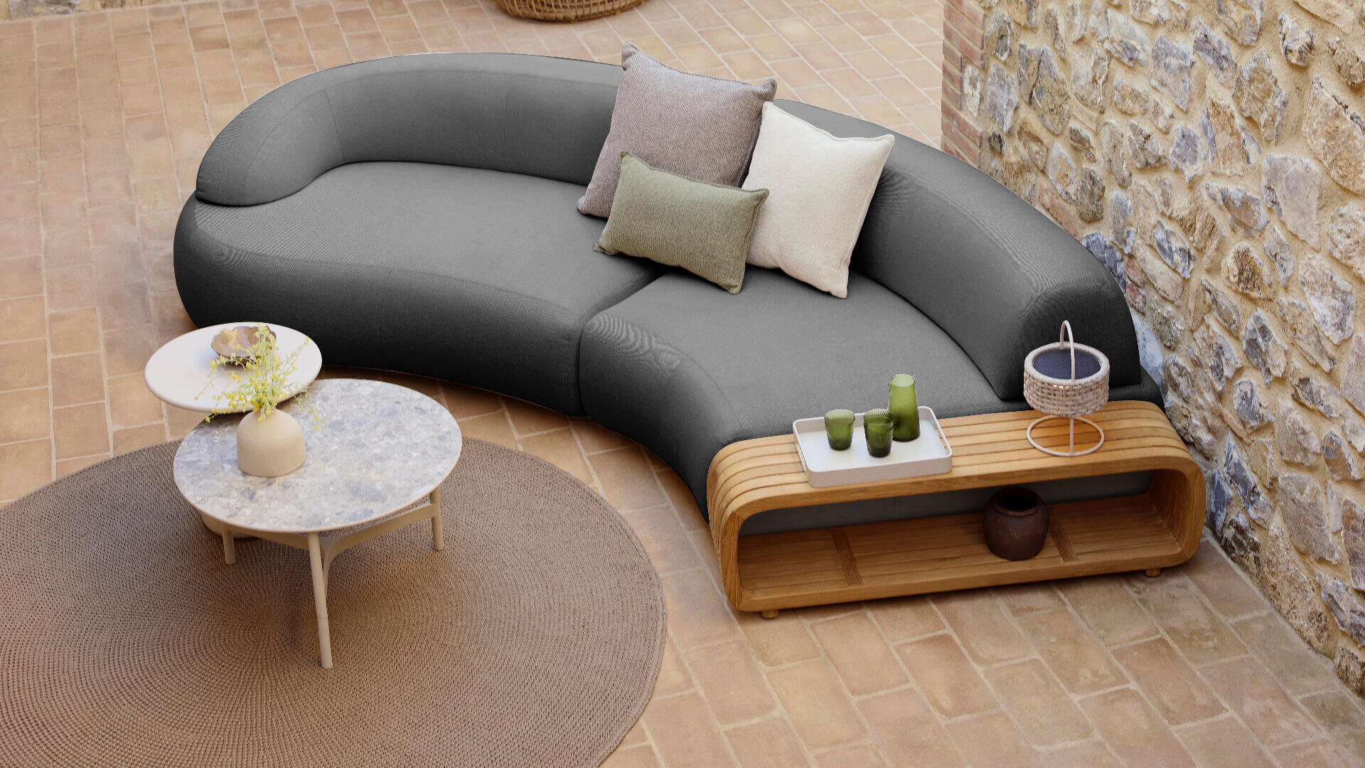 Curved gray sofa with four decorative pillows, next to a wooden side table holding green cups, a basket, and a candle, on a tan tiled floor with a round beige rug and round marble coffee table. Stone wall in the background.
