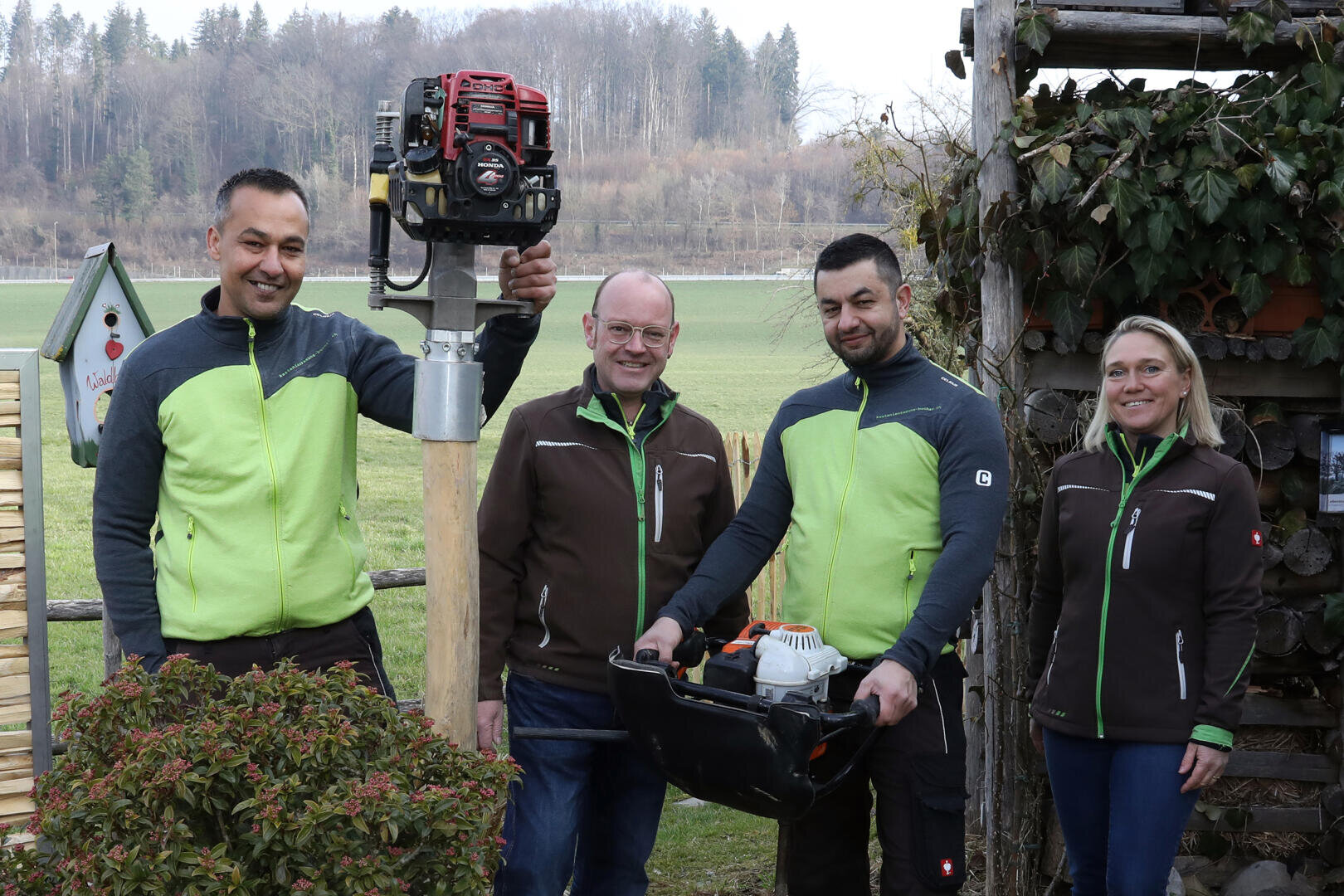 Vier Personen posieren im Freien in grünen und braunen Arbeitsjacken. Zwei Männer halten Werkzeuge, darunter ein Stangenwerkzeug und einen schwarzen Koffer. Im Hintergrund sind Felder und Bäume zu sehen, in der Nähe befinden sich Holzkonstruktionen und Pflanzen.