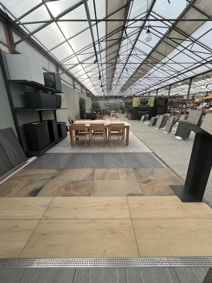 In einem geräumigen Ausstellungsraum mit einer Glasdecke werden verschiedene Bodenfliesen ausgestellt. In der Mitte steht ein Esstisch aus Holz mit Stühlen, umgeben von Fliesenmustern und großen Regalen an den Wänden.