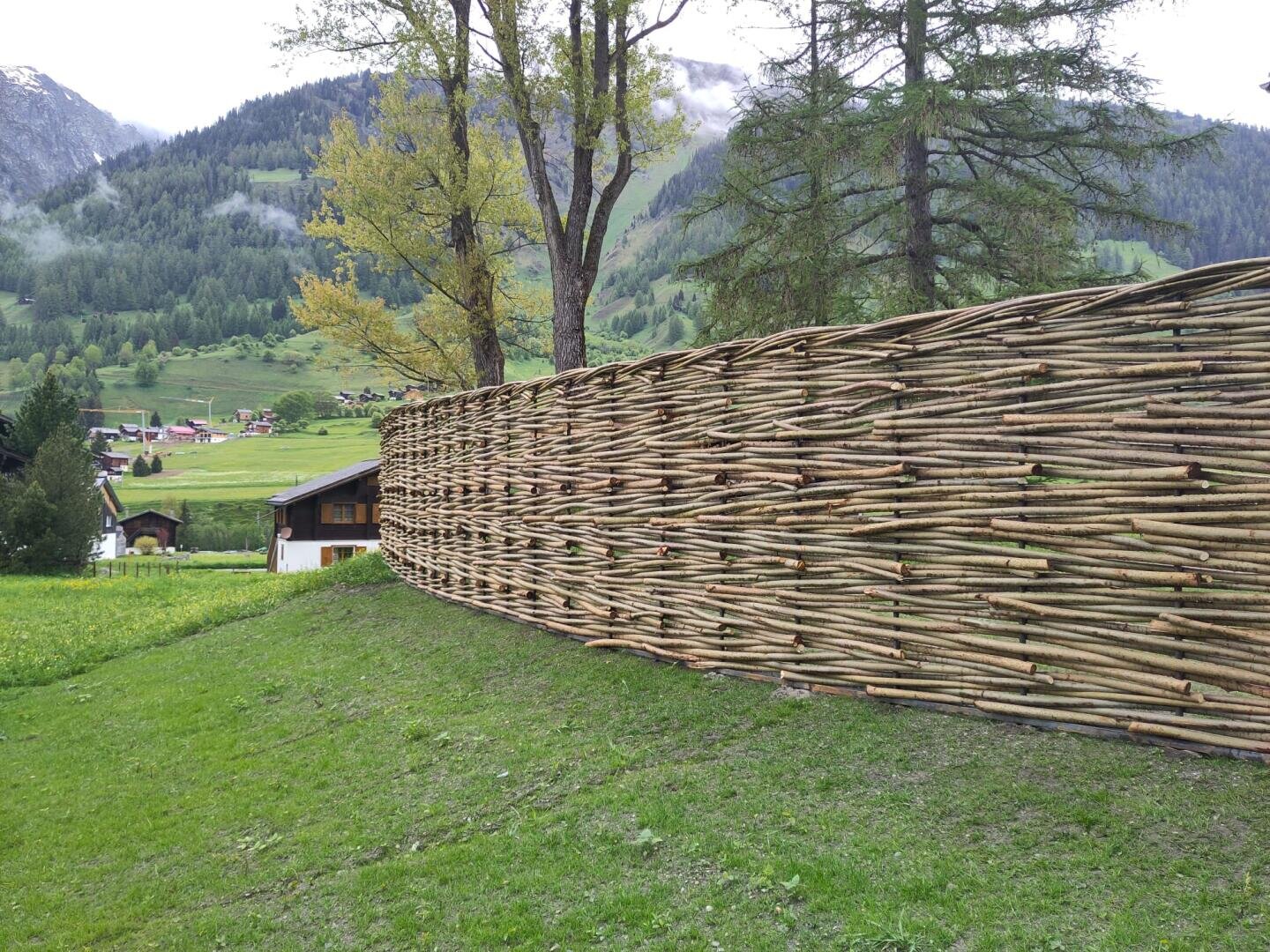 A tall woven fence made of thin wooden branches stands on a grassy slope, with trees, hills and a small house in the background under a cloudy sky.