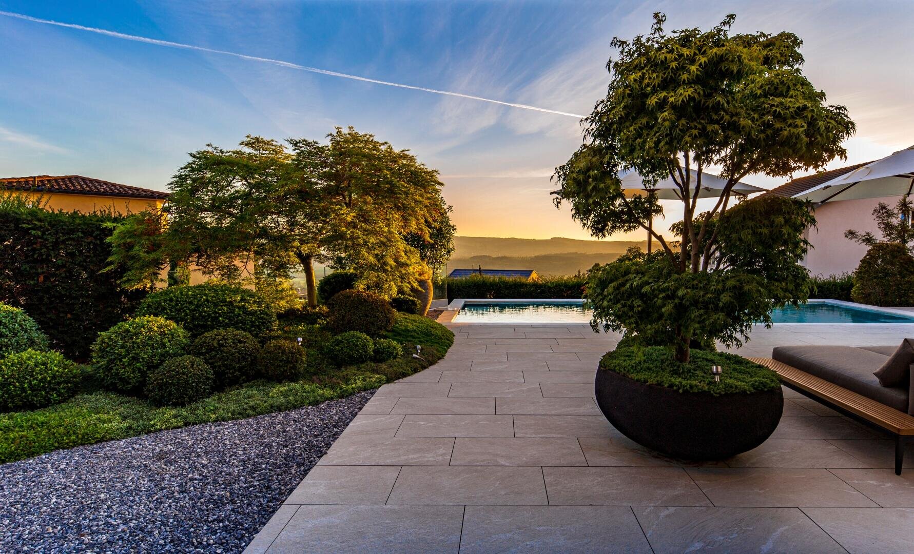 Ein Garten bei Sonnenuntergang mit einem Baum in einem großen Pflanzgefäß, gepflegten Sträuchern, Steinfliesen, Kies und einem Pool mit Liegestühlen, mit Blick auf ferne Hügel unter einem klaren Himmel.