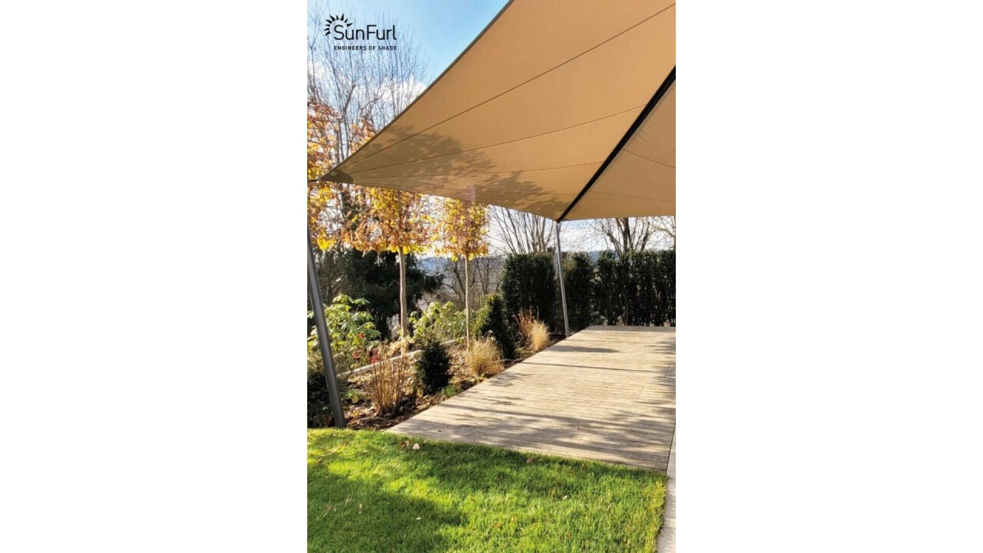 A triangular beige shade sail is stretched over a wooden deck in a garden, with green grass, shrubs, and trees visible under a sunny sky. The SunFurl company logo appears at the top left corner.