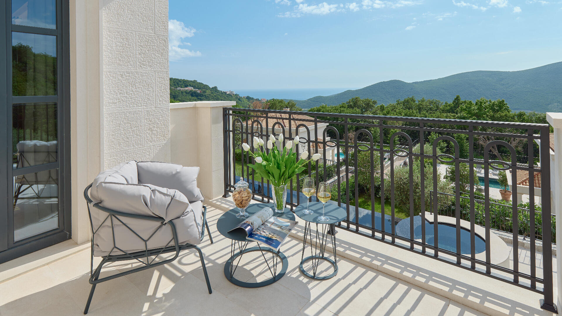 Ein sonniger Balkon mit einem gepolsterten Stuhl, zwei kleinen Tischen mit einer Blumenvase, einem Buch, einer Sonnenbrille und Getränken. Der Blick schweift über grüne Hügel, Dächer und das ferne Meer unter blauem Himmel.