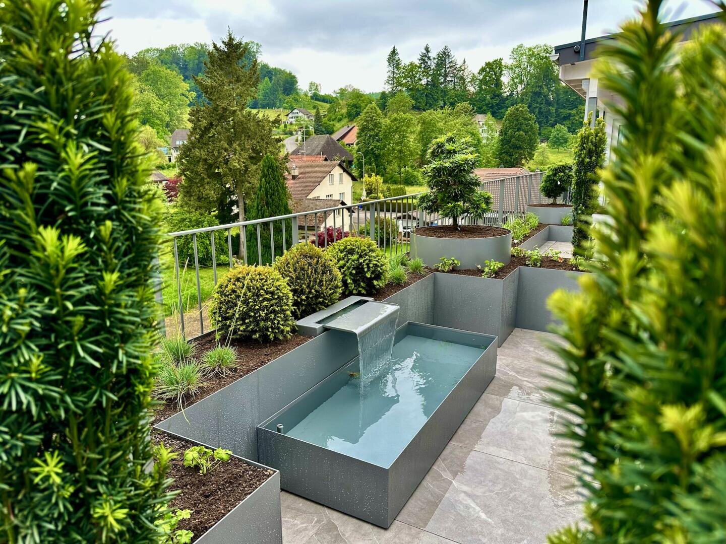 Ein moderner Terrassengarten mit ordentlich geschnittenen Sträuchern, erhöhten Pflanzgefäßen und einem rechteckigen Wasserspiel mit einem metallischen Ausguss, mit Blick auf eine üppige, grüne Landschaft und entfernte Häuser.