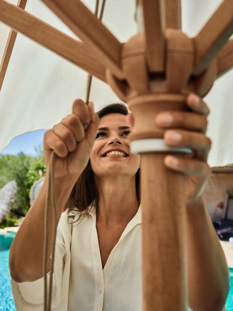 A woman in a white shirt smiles as she raises or adjusts a large wooden patio umbrella by a pool, with greenery and a sunlit outdoor setting in the background.