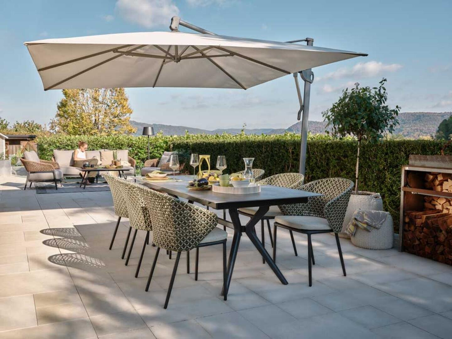 Moderne Außenterrasse mit einem Esstisch für sechs Personen unter einem großen weißen Sonnenschirm, umgeben von Grünpflanzen und Loungesitzen im Hintergrund, an einem sonnigen Tag.