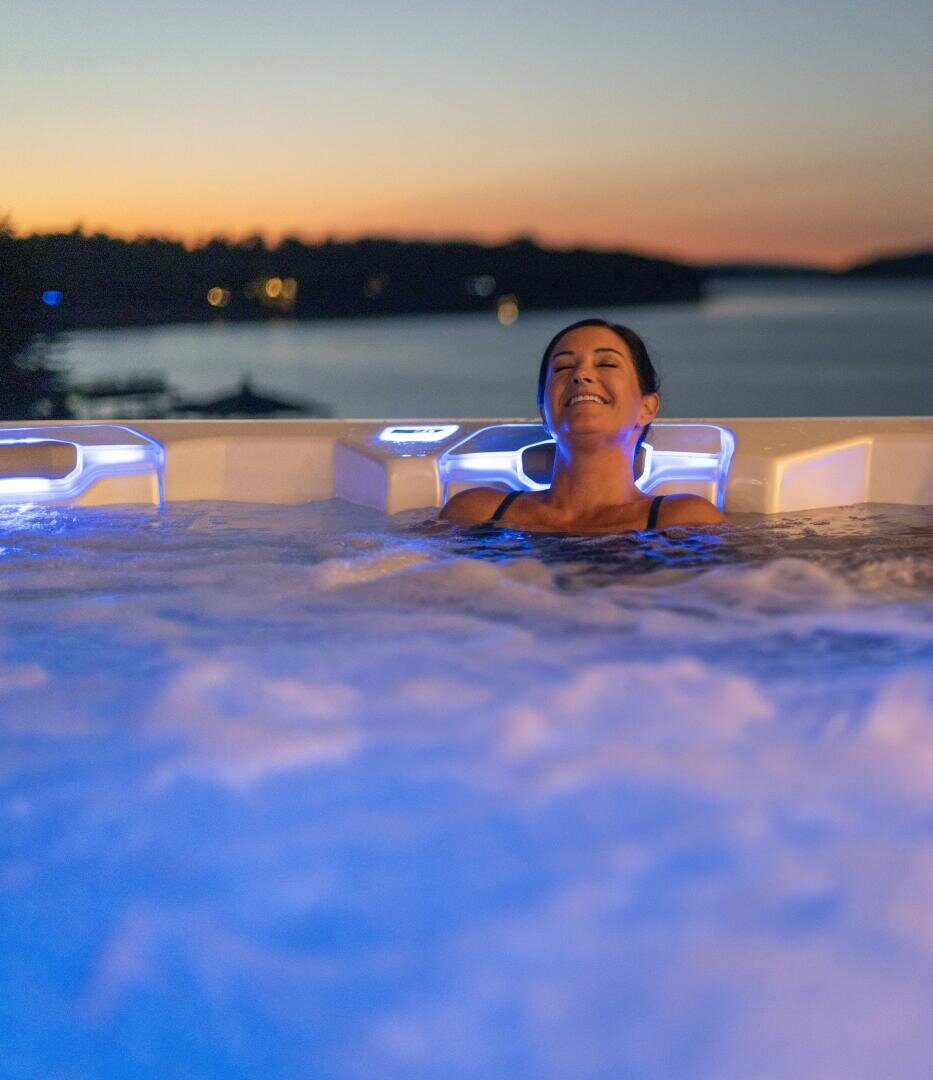 Eine Person entspannt sich in einem Whirlpool mit blauen Lichtern, die Augen geschlossen und lächelnd, während die Sonne über einem ruhigen See und silhouettierten Bäumen im Hintergrund untergeht.