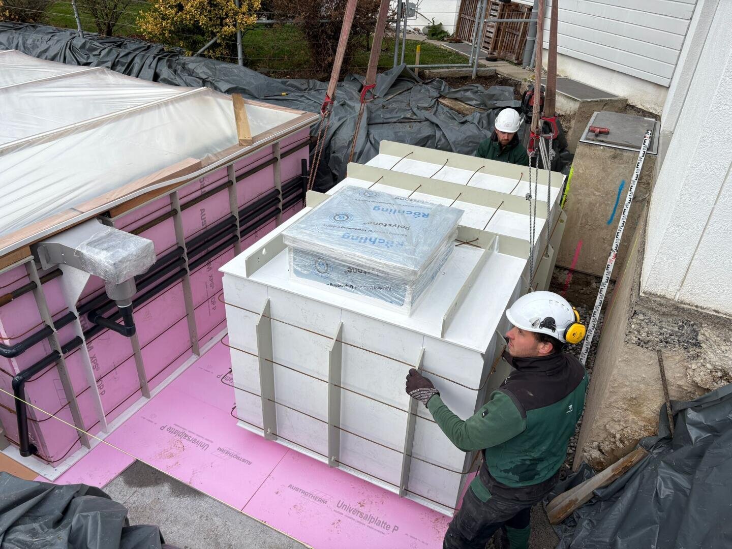 Zwei Bauarbeiter mit Helmen führen ein großes, weißes Fertigteil, das auf einer rosa Schaumstoffunterlage auf einer Baustelle im Freien neben einem weißen Gebäude abgesetzt wird.