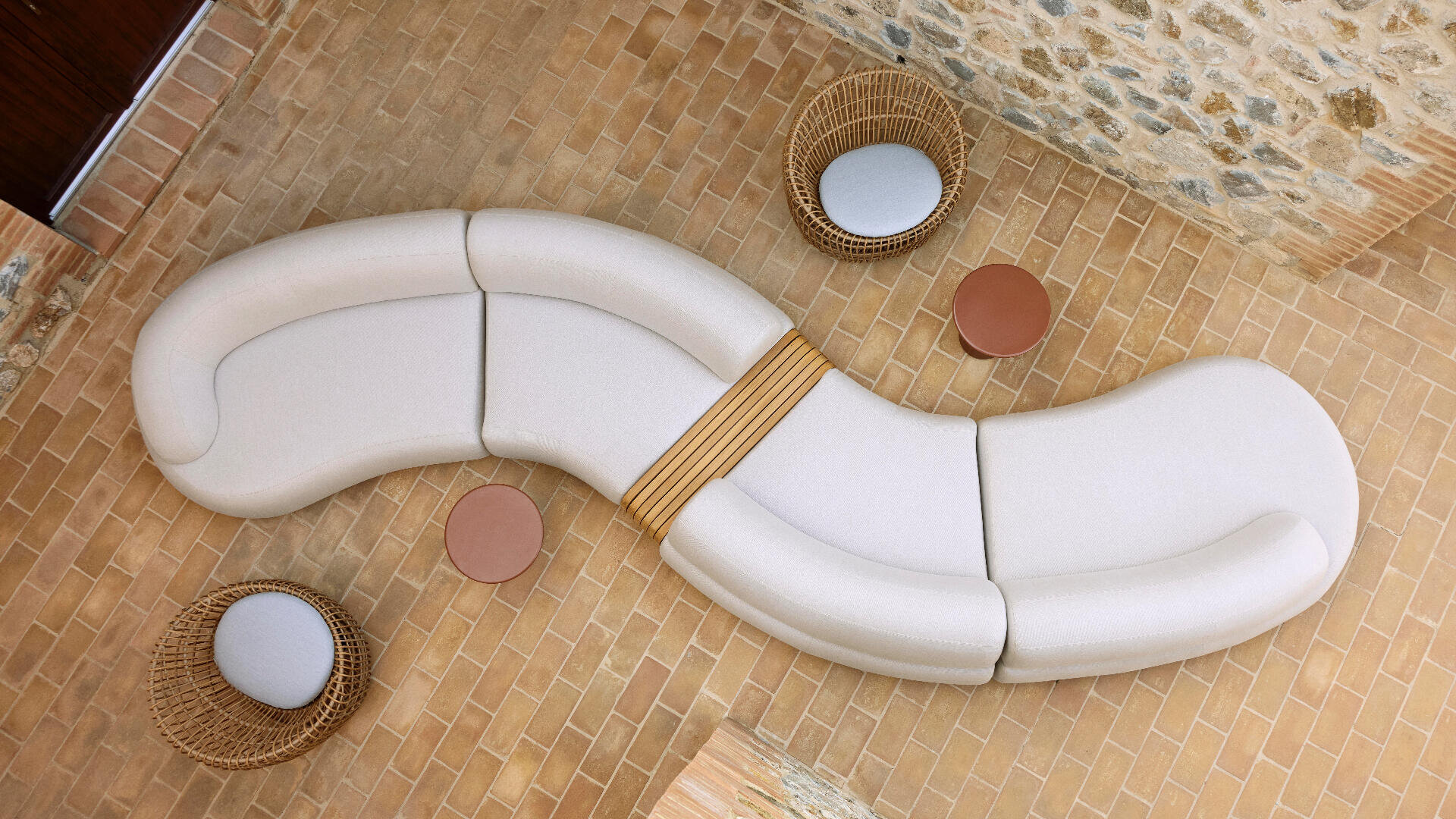 Curved white sectional sofa on tan brick floor, with two circular wicker chairs, two round tan side tables, and stone wall in background, viewed from above.
