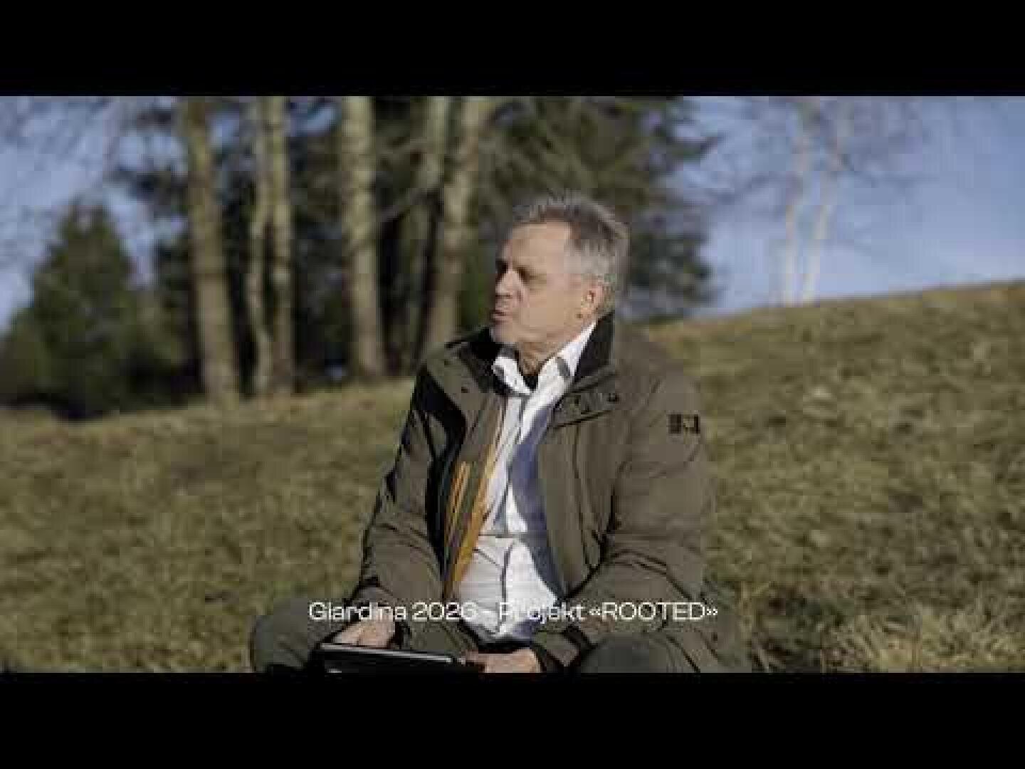 Ein Mann in einer grünen Jacke sitzt im Freien auf einem grasbewachsenen Hang und hält ein Tablet in der Hand. Im Hintergrund sind Bäume und blauer Himmel zu sehen. Der Text am unteren Rand lautet: Glarolna 2023, Projekt "ROOTED".